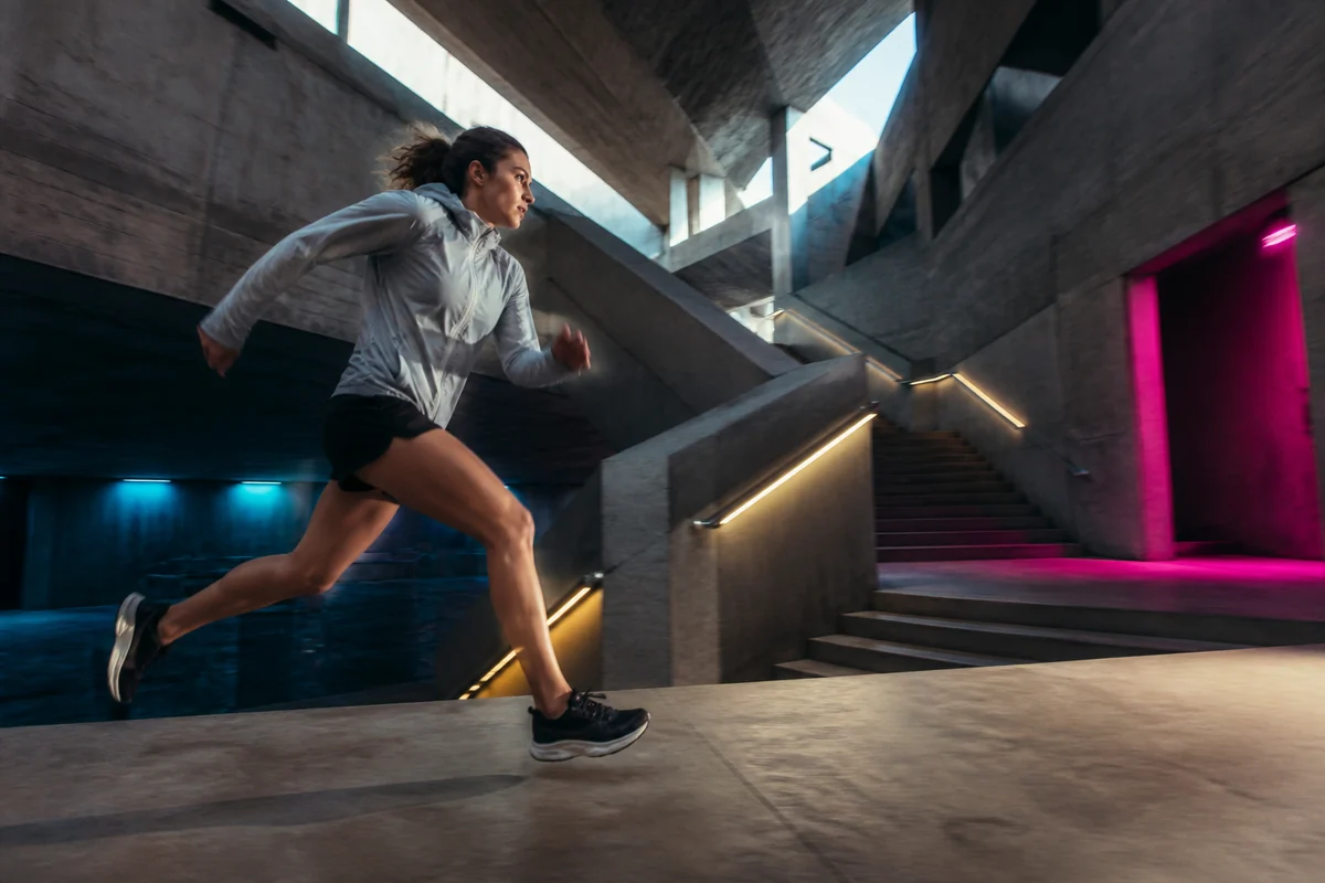 Eine Sportlerin läuft durch ein brutalistisches Treppenhaus, dessen Licht und Architektur ihren Weg sichtbar lenken.