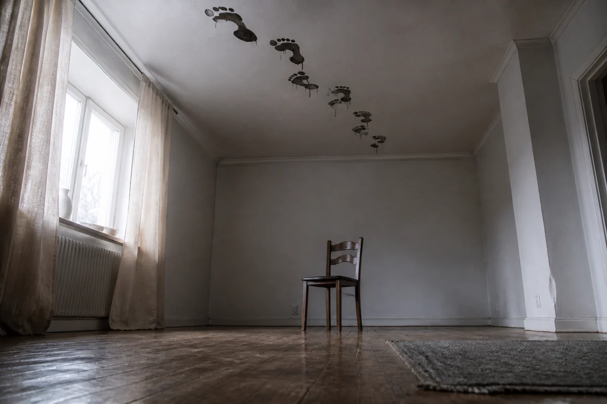 Nasse schwarze Fußabdrücke laufen in einem hellen leeren Zimmer über die Decke und enden über einem verrückten Holzstuhl.
