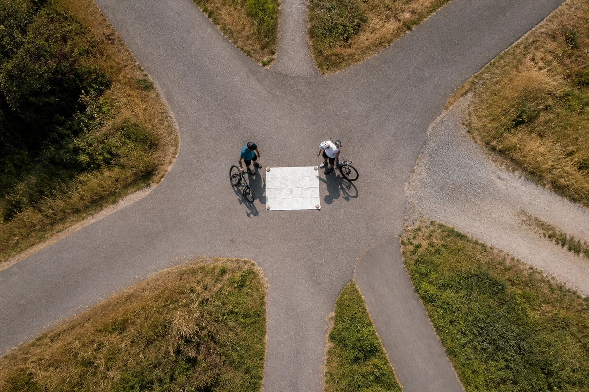 Zwei Radfahrer halten aus senkrechter Vogelperspektive an einer komplexen Weggabelung neben einer großen neutralen Papierkarte.